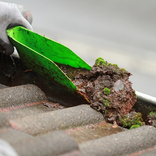 A green scoop removing muck from a clogged gutter.